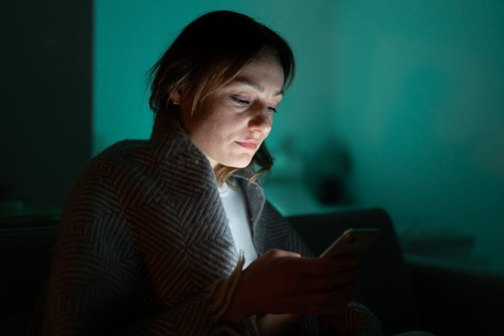 Young woman sitting in the dark scrolling through her phone.