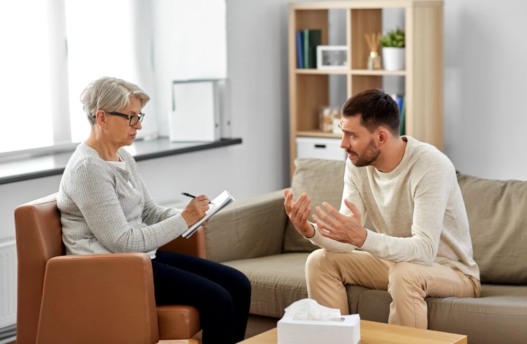 Young man talking to a therapist, who is writing down his symptoms in a notebook.