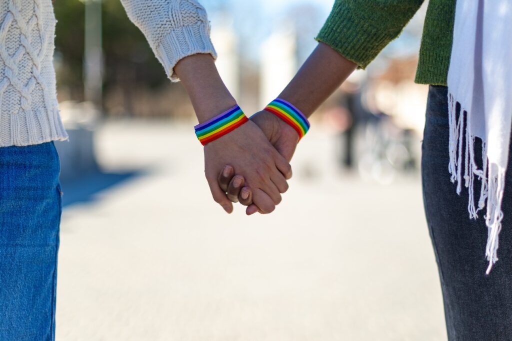 Two LGBTQ individuals holding hands with Pride flag bracelets.