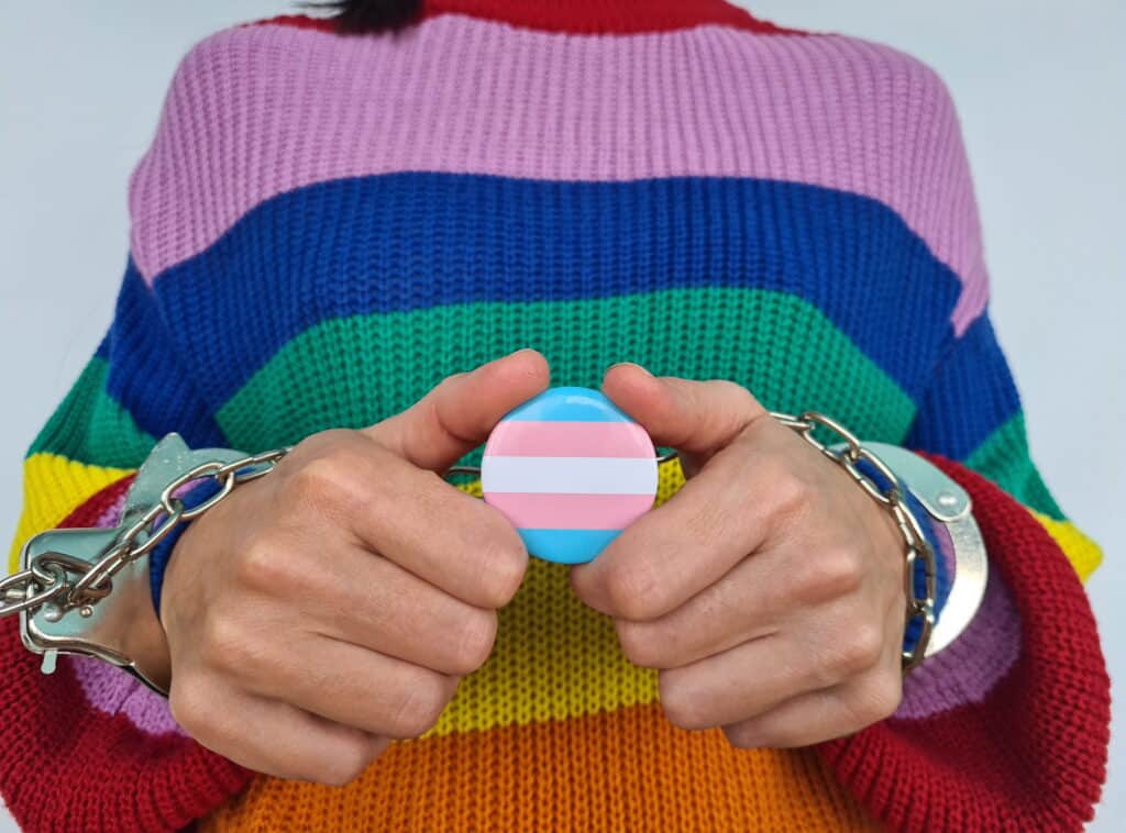Individual wearing a rainbow sweater and holding a transgender flag pin.
